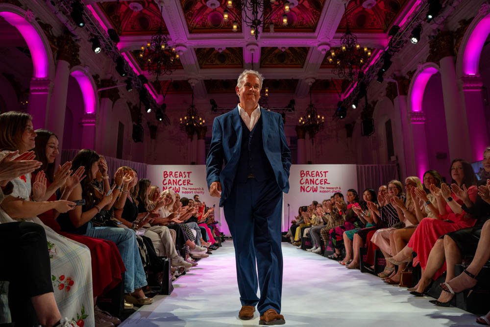 Dave Talbot walking the runway in a blue three-piece suit at Breast Cancer Now's The Show 2025, applauded by the audience under pink lights